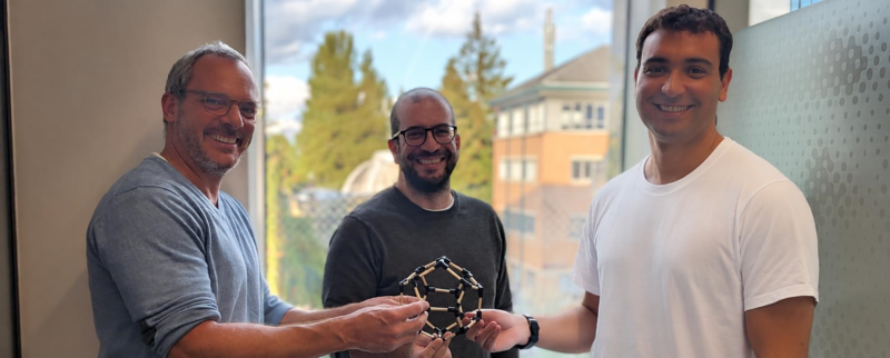 Three men smile while holding a molecular model in an office with a window view in the background.