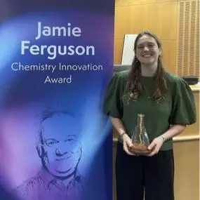 A smiling woman holds a trophy beside a banner for the Jamie Ferguson Chemistry Innovation Award at the University of Oxford Department of Chemistry.