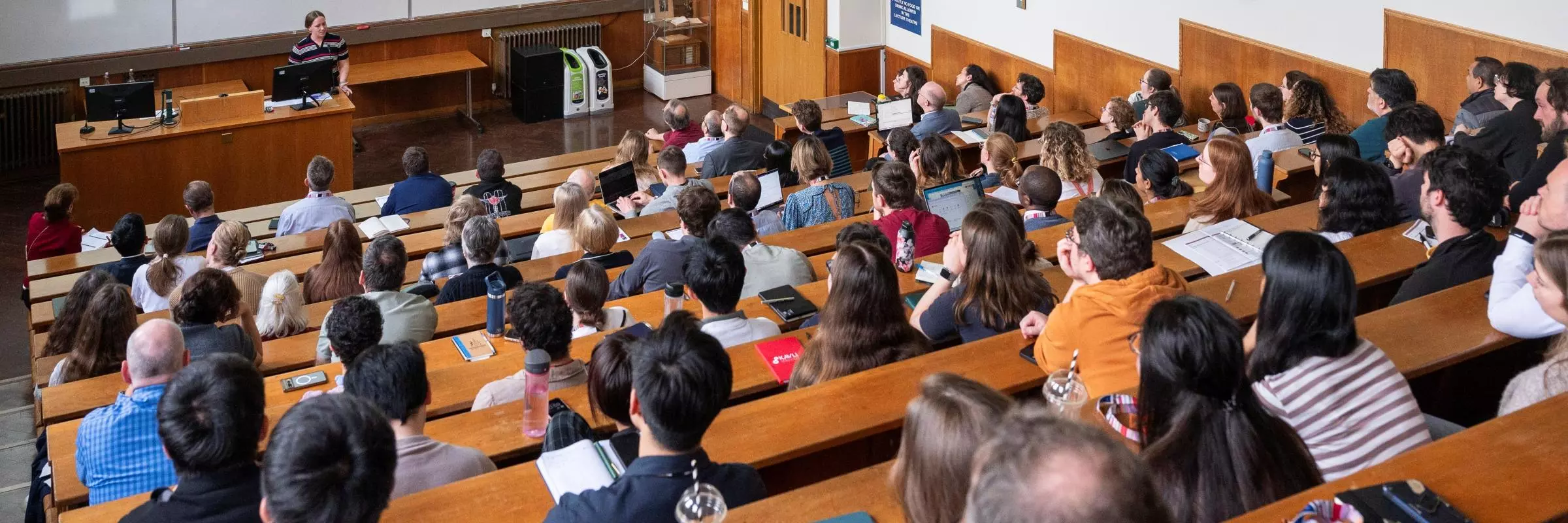 A large audience attends a lecture in a tiered university lecture theatre, listening to a presenter at the front. Many attendees have laptops, notebooks and papers on the wooden desks.